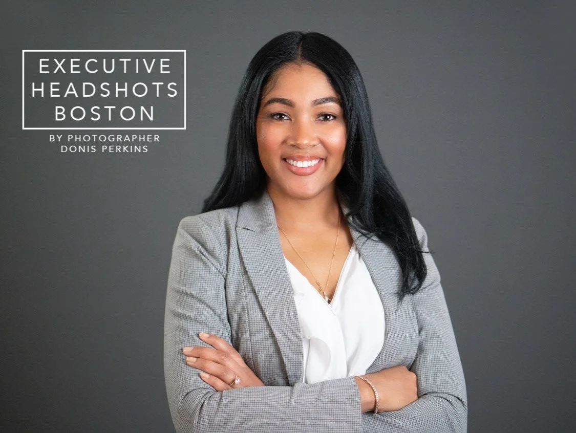 Smiling woman with long black hair in a grey checked blazer over a white blouse, arms folded, on a dark grey background