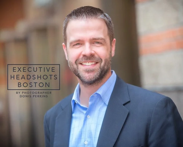 Smiling man with a beard in a navy blazer over a blue checked shirt against a brick wall