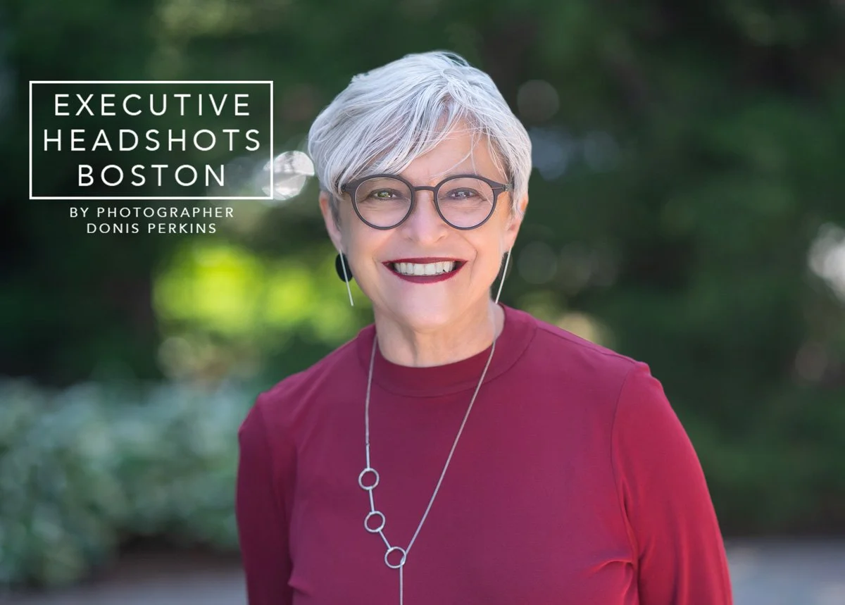 Smiling woman with short white hair and round glasses in a deep red top and long silver necklace, outdoors among greenery