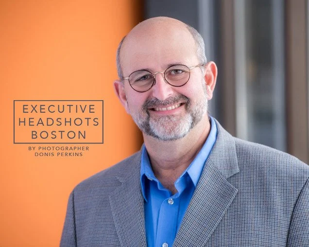 Smiling man with a grey beard and round glasses in a grey checked jacket over a blue shirt beside an orange wall